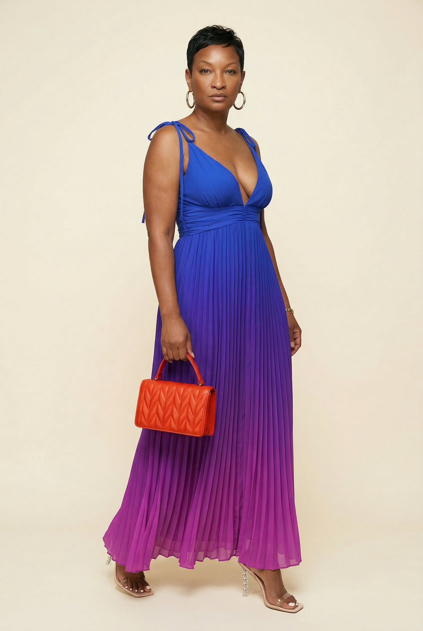 Woman wearing a blue and purple pleated dress holding an orange handbag on a beige background